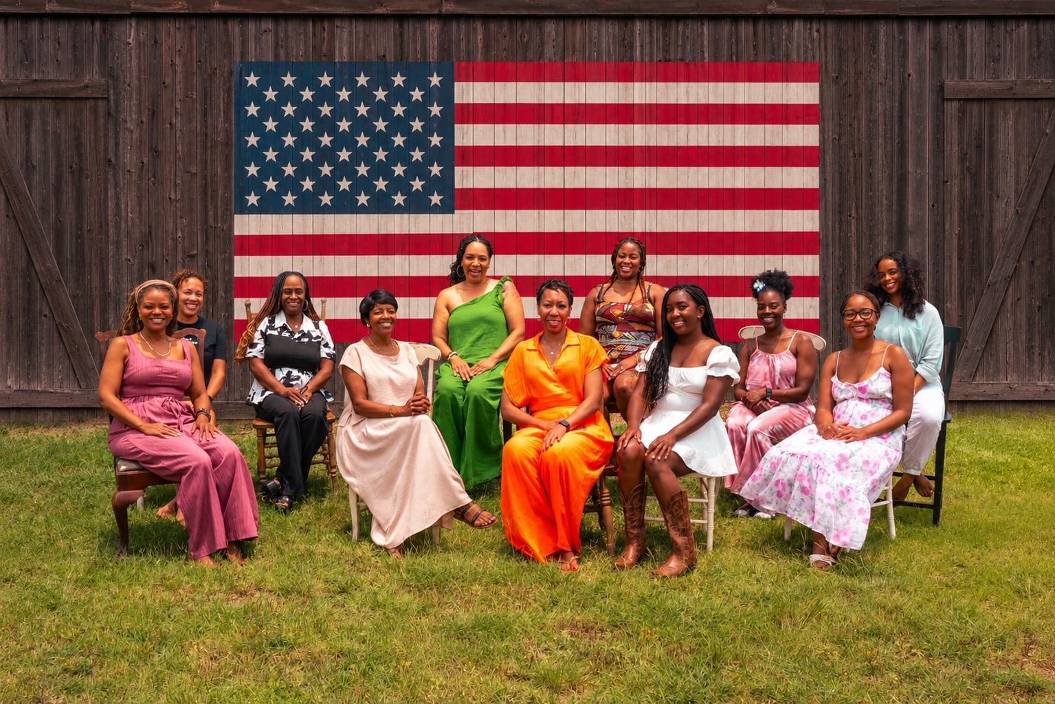 Founders photo. Well-rested Black women on Juneteenth, seated together in front of a wooden barn wall with a hand-painted American flag mural.