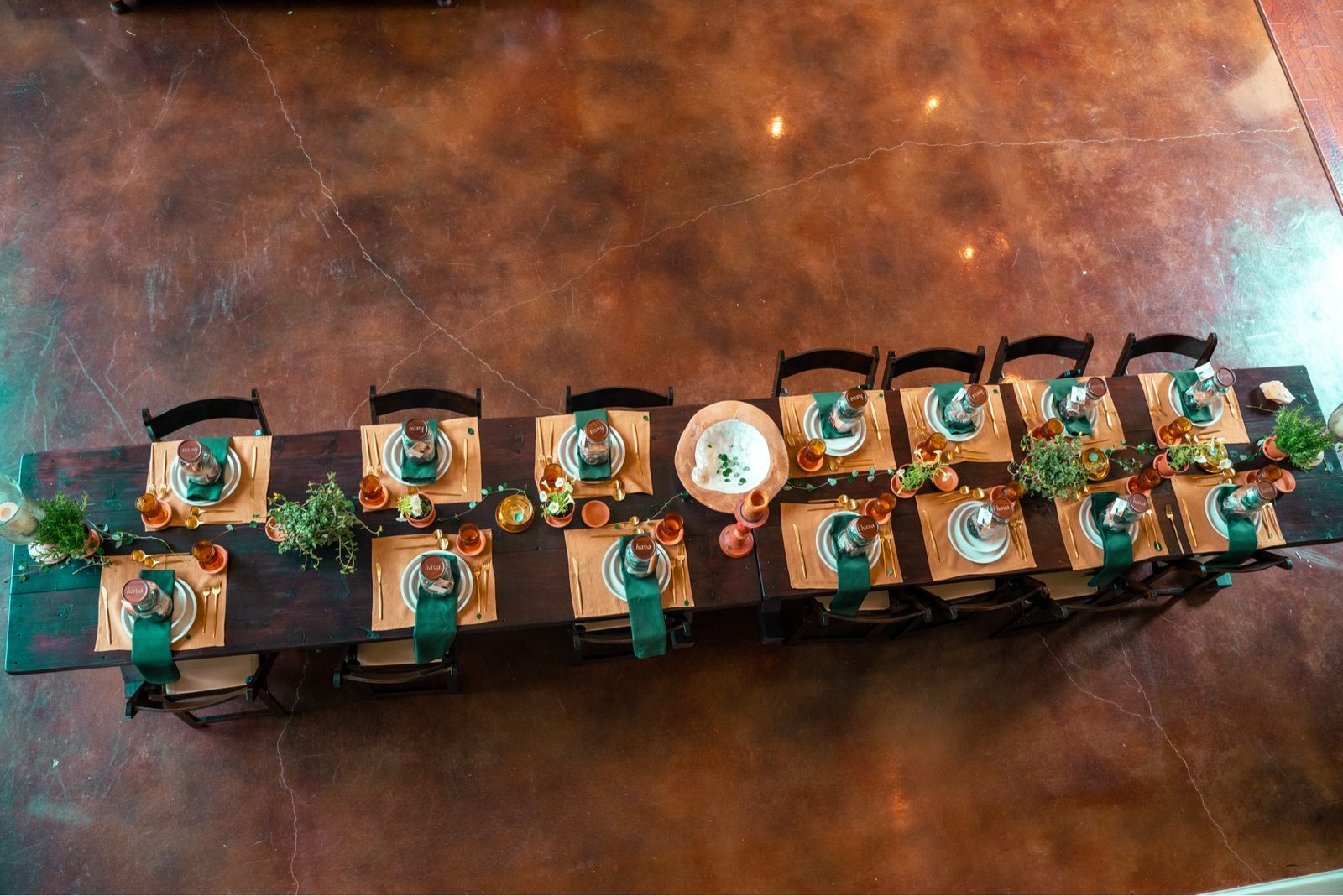 A long communal table set with green napkins, gold flatware, terracotta place settings, and greenery, viewed from above.