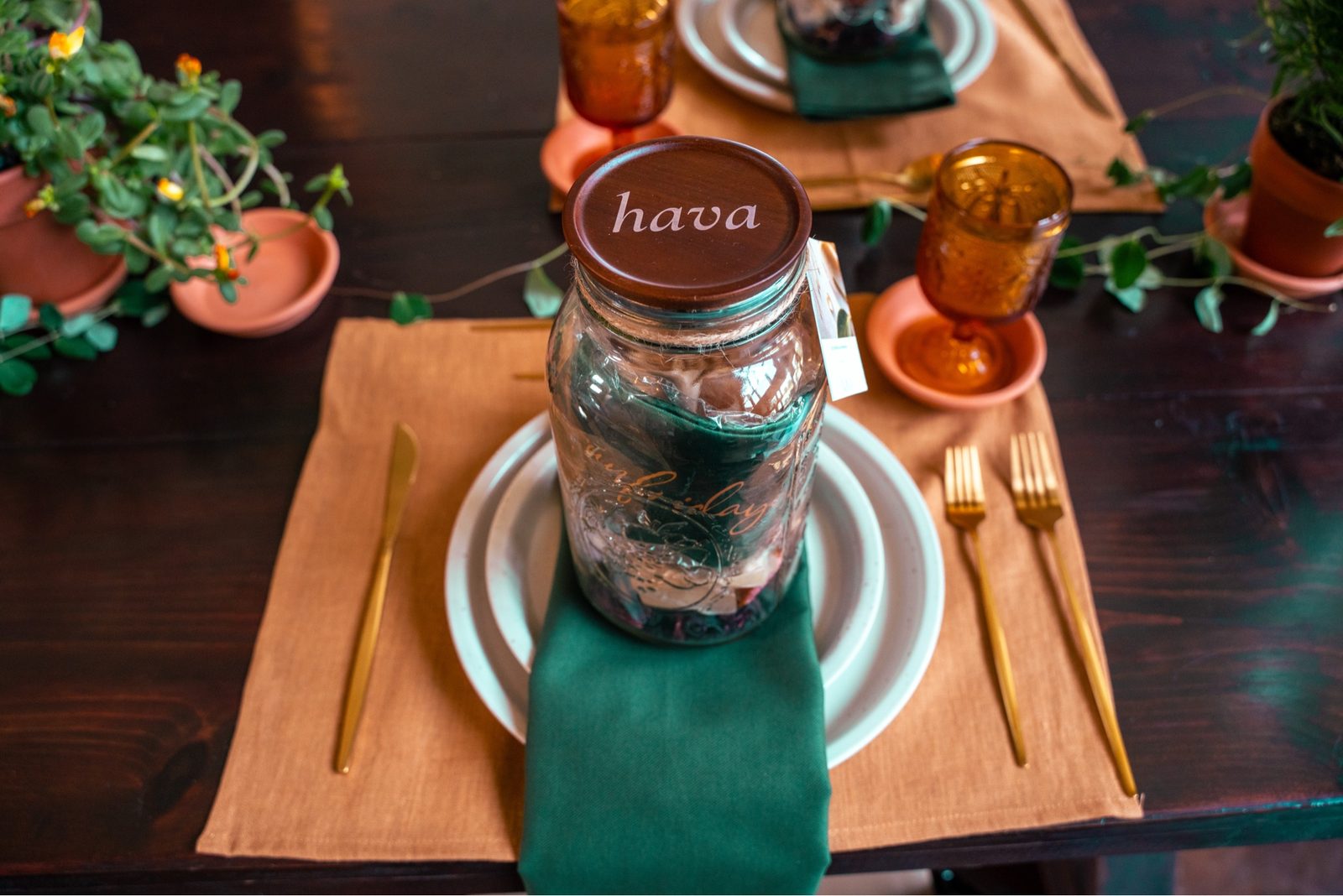 A place setting close-up: a mason jar with a hava wood lid rests on a white plate wrapped with a green napkin, gold cutlery beside.
