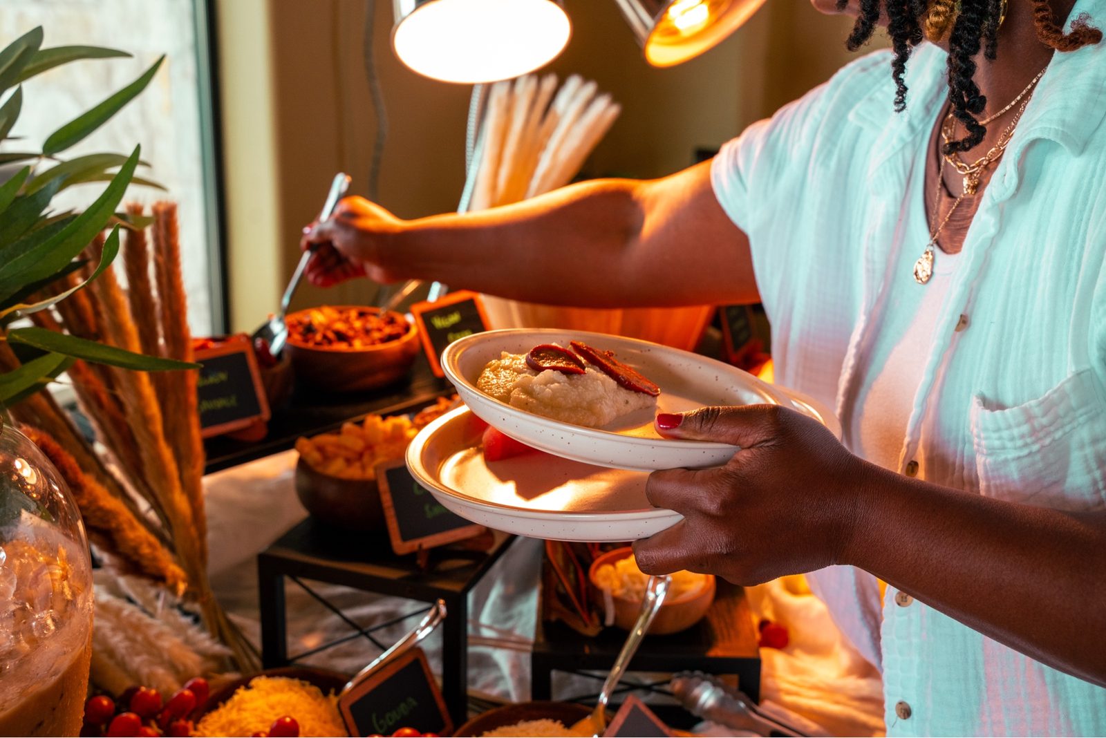 A woman in mint blue makes a plate at the buffet, warm heat lamps overhead.