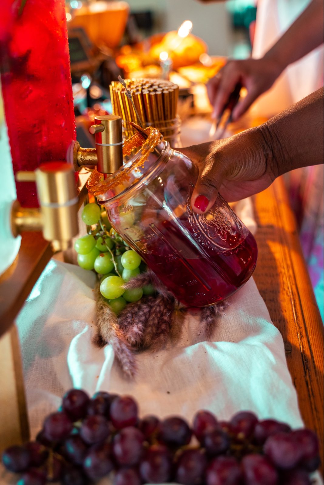 A woman's hands pour a deep red drink from a spigot into a mason jar, grapes and gold reeds arranged below.