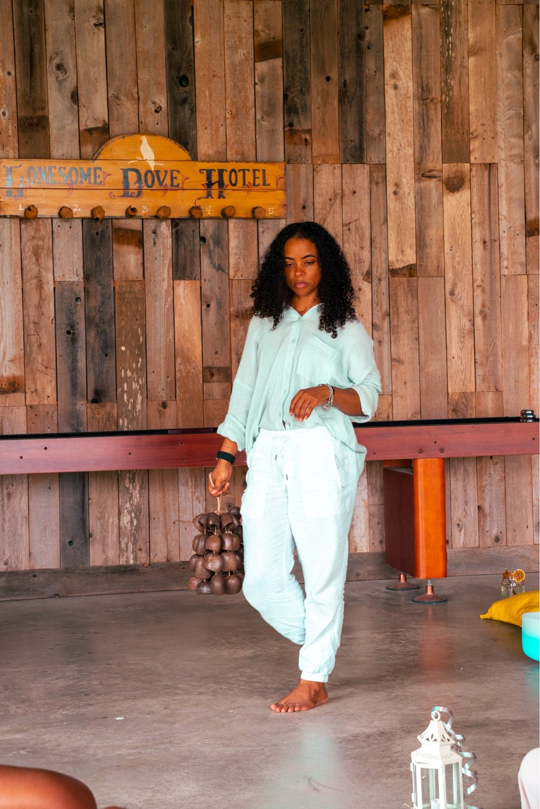 A woman in a mint linen set holds a cluster of clay bells before a wood-plank wall bearing a Lonesome Dove Hotel sign.