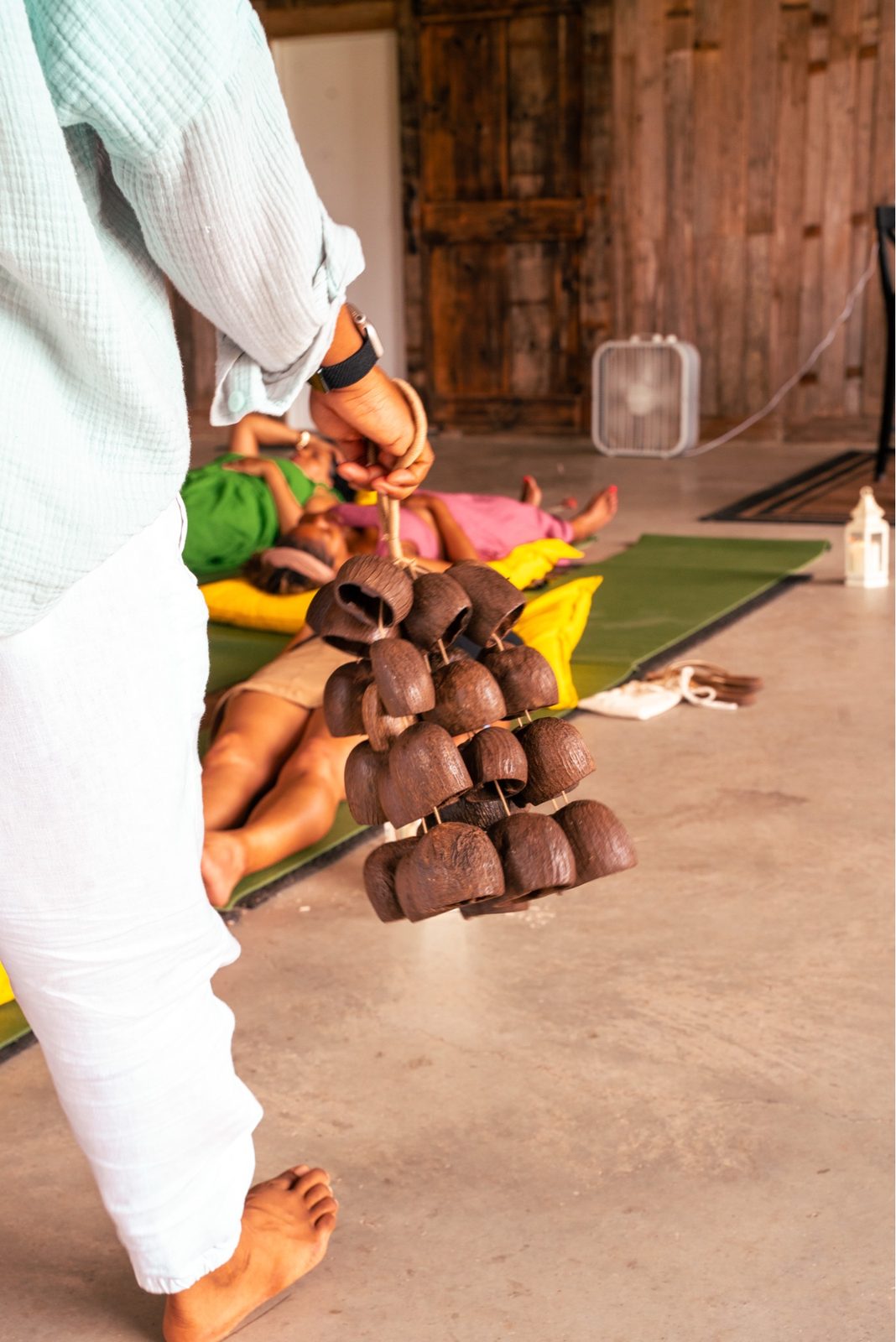 Women rest on yoga mats in savasana, a hand in the foreground holds a cluster of clay bells.