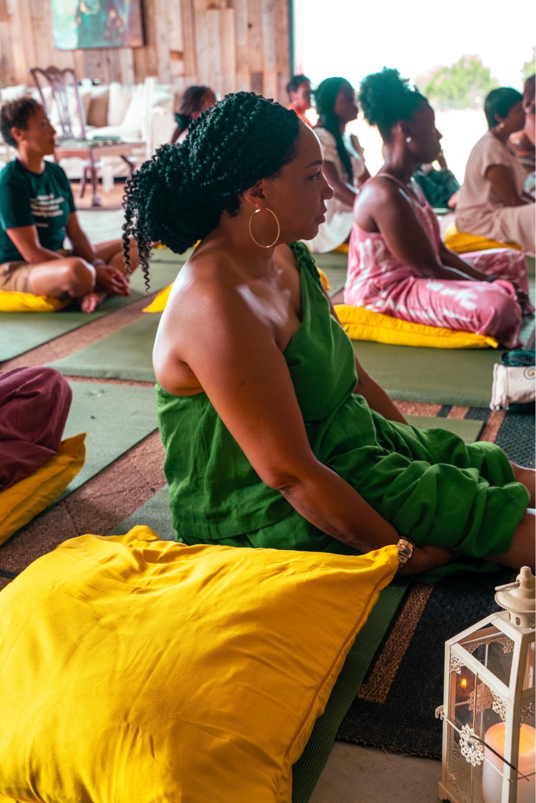 A woman in a green linen set sits cross-legged on a mat in sister circle, hoop earring visible, other women seated around her.