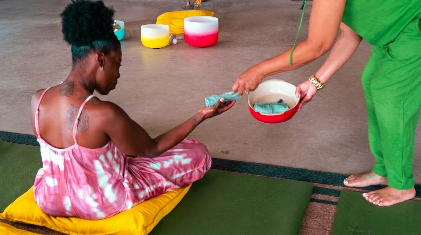 A woman receives a folded cloth from another woman's hand over a red bowl, crystal singing bowls arranged on the floor behind her.