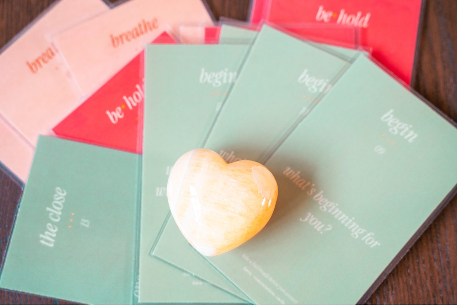Practice cards fanned on a table reading breathe, be·hold, begin, the close — with a smooth honey-colored heart-shaped stone resting on top.