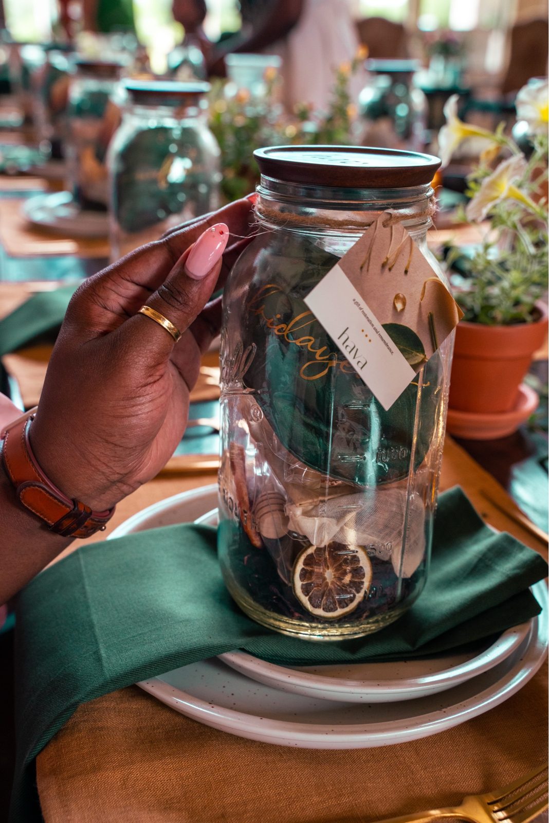A woman with a gold ring holds up a hava mason jar keepsake, a paper tag hanging from the lid, dried orange slice inside.